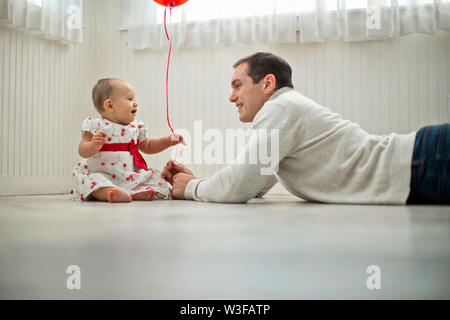 Giovane padre giocando sul pavimento con la sua bambina e un galleggiante rosso palloncino che egli ha dato la sua. Foto Stock