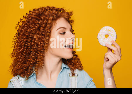 Ricci i capelli rossi donna ciambella azienda desidera mangiare ciambella dolce su sfondo giallo Foto Stock