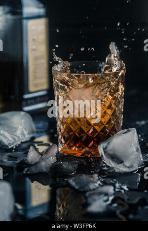 Alcoliche bere whisky in un bicchiere di ghiaccio con una bottiglia in background, su uno sfondo nero, close-up con schizzi Foto Stock