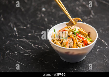 Tagliatelle cinesi in una tazza con il pollo e le verdure con bastoni. Wok cibo dal ristorante su sfondo scuro da una vista laterale. Foto Stock