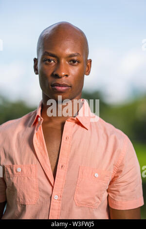 Ritratto di un bel giovane americano africano maschio modello guardando la fotocamera. Uomo calvo che indossa un pulsante rosa shirt su sfondo sfocato Foto Stock