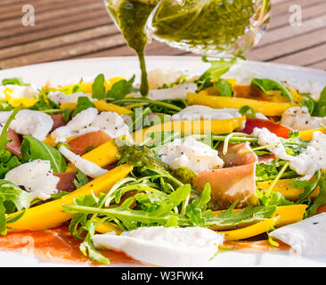 Estate cibo: insalata pasto servito all'aperto con fette di mango, rucola, mozzarella e prosciutto di Parma sulla piastra bianca con basilico medicazione versata sopra Foto Stock