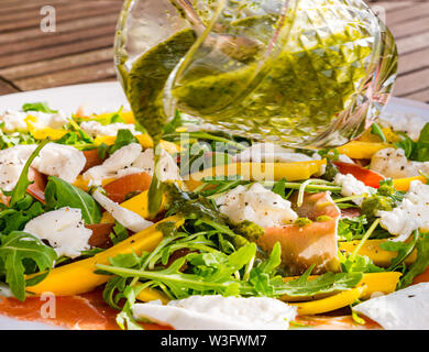 Estate cibo: insalata pasto servito all'aperto con fette di mango, rucola, mozzarella e prosciutto di Parma sulla piastra bianca con basilico medicazione versata sopra Foto Stock