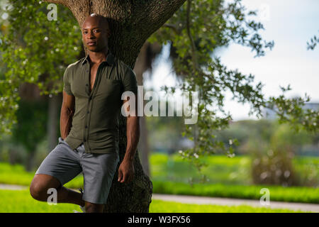 Bello rilassato uomo appoggiato a un albero nel parco. African American modello indossando una maglietta stretta che mostra i muscoli off Foto Stock