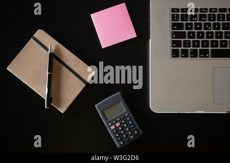 Vista parziale di un argento della tastiera di un portatile con una calcolatrice, una rosa post-it e un notebook con una matita su una scrivania di legno scuro Foto Stock