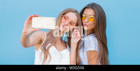 Moda ritratto di due graziose hipsters sorridente donna in occhiali da sole tenendo lo smartphone e rendendo selfie contro la coloratissima parete blu. Foto Stock