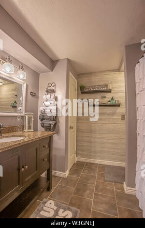 Bagno con specchio vanity rivolta verso la vasca da bagno e doccia dietro la tenda bianca. Porta asciugamani e mensole sono montati sulla parete grigia. Foto Stock