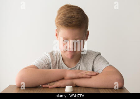 Rosso-testa ragazzo caucasico siede a un tavolo con un unico marshmallow, tentare la prova di marshmallow Foto Stock