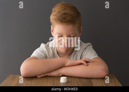 Rosso-testa ragazzo caucasico siede a un tavolo con un unico marshmallow, tentare la prova di marshmallow Foto Stock