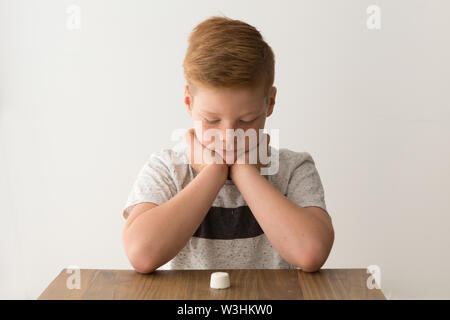 Rosso-testa ragazzo caucasico siede a un tavolo con un unico marshmallow, tentare la prova di marshmallow Foto Stock