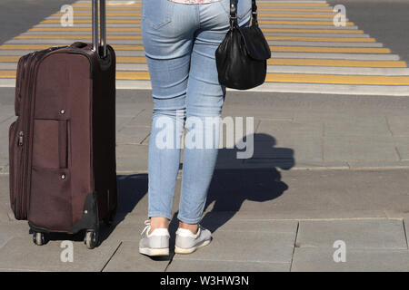 Donna con una valigia su ruote sorge su un attraversamento pedonale, in vista posteriore Gambe femmina in jeans blu sul crosswalk, viaggi, taxi in attesa Foto Stock