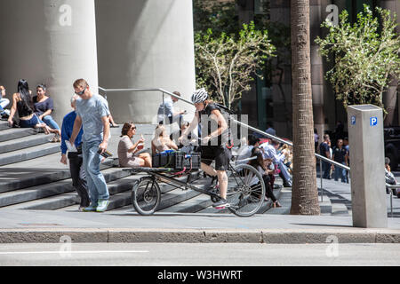 Bicicletta peddles corriere in salita nel centro cittadino di Sydney passato impiegati godendo il pranzo sui passi di n. 1 Bligh Street, Sydney, Australia Foto Stock