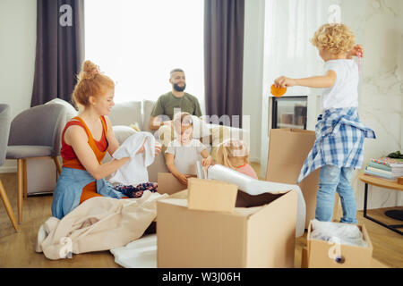 Tanto lavoro. Nice positivo i bambini seduti intorno le caselle mentre aiuta la loro madre di spacchettarli Foto Stock