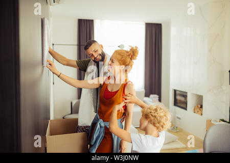 Metterla lì. Piacevole ragazzo biondo aiutando i suoi genitori mentre dando loro consigli dove mettere la pittura Foto Stock