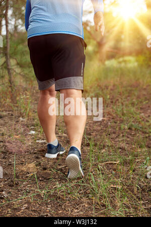 Uomo adulto in vestiti blu e pantaloncini nero corre lungo la foresta di conifere contro il sole luminoso, il concetto di uno stile di vita sano e in esecuzione in Foto Stock