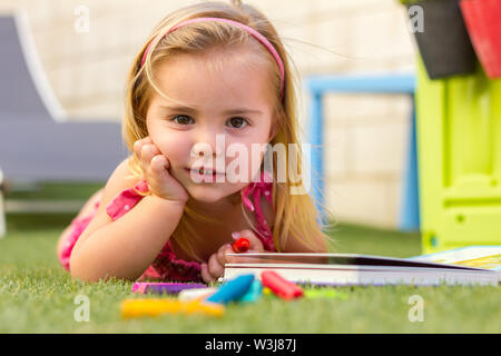 Ritratto di un simpatico bionda bambina posa sull'erba. Ella è la lettura di un libro e ci sono alcuni pastelli a cera sul pavimento Foto Stock