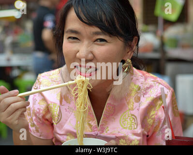 Vestite Thai donna cinese con naso rugoso mangia giallo tagliatelle con bacchette durante il Nuovo Anno Cinese. Foto Stock
