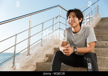 Sorridente giovane sportivo asiatico seduto sulle scale in riva al mare Foto Stock