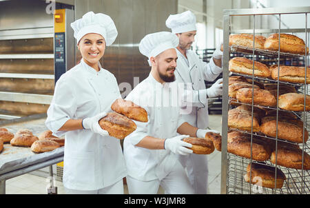 Un baker donna trattiene il pane fresco con la sua mano contro lo sfondo di fornai a lavorare in una panetteria. Foto Stock