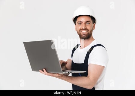 Fiducioso felice barbuto builder uomo indossare tuta e casco in piedi isolato su sfondo bianco, utilizzando il computer portatile Foto Stock