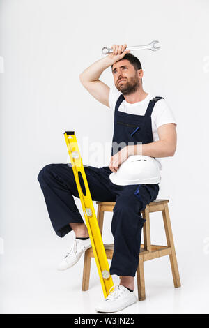 Per tutta la lunghezza di uno stanco barbuto builder uomo indossare tute e hardhat seduto su una sedia isolate su sfondo bianco Foto Stock