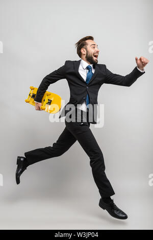 Lunghezza completa di un attraente sorridente giovane imprenditore indossare tuta isolate su uno sfondo grigio, portando sullo skateboard, in esecuzione Foto Stock