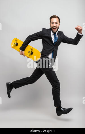 Lunghezza completa di un attraente sorridente giovane imprenditore indossare tuta isolate su uno sfondo grigio, portando sullo skateboard, in esecuzione Foto Stock