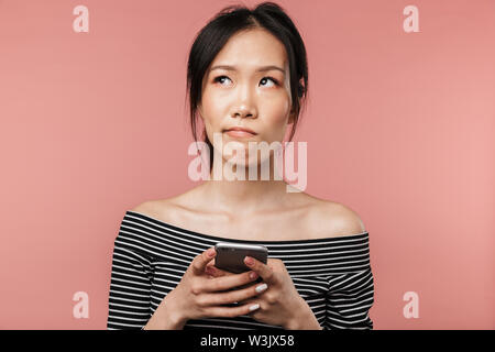 Foto di giovane donna asiatica vestita di base del pensiero di usura e cercando in disparte mentre si tiene lo smartphone isolate su sfondo rosso in studio Foto Stock