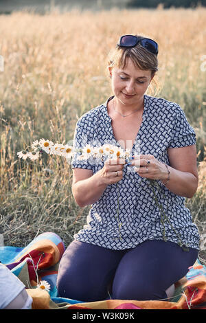Sorridendo felice donna fare coronet di fiori selvatici. Famiglia di trascorrere del tempo insieme su un prato, vicino alla natura. Candide persone, momenti di vera e autentica Foto Stock