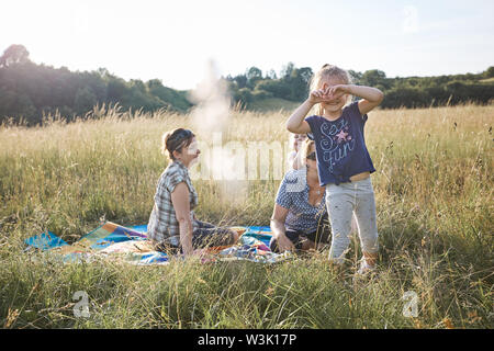 Bambina facendo un gesto a mano per scattare la foto. Famiglia di trascorrere del tempo insieme su un prato, vicino alla natura. I genitori e i bambini seduti su un blanke Foto Stock