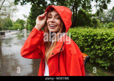 Immagine del Caucaso in giovane donna 20s in un colorato impermeabile avente divertimento mentre passeggiate attraverso il parco verde sotto la pioggia Foto Stock