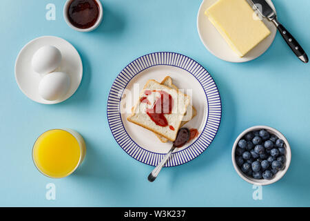 Vista dall'alto di fette biscottate con marmellata, succo d'arancia, uova sode, burro e mirtilli su sfondo blu Foto Stock