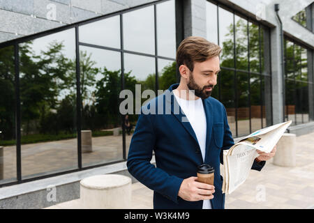 Ritratto di imprenditore fiducioso nella camicia di bere il caffè dal bicchiere di carta e giornale di lettura permanente, mentre all'esterno vicino all'edificio Foto Stock