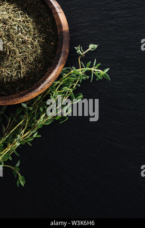 Vista superiore del timo essiccato in ciotola di legno vicino a erba verde su sfondo nero Foto Stock