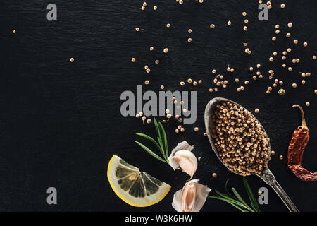 Vista dall'alto di semi di coriandolo in cucchiaio d'Argento su nero testurizzato sfondo con peperoncino essiccato pepe, limone, erbe e aglio Foto Stock