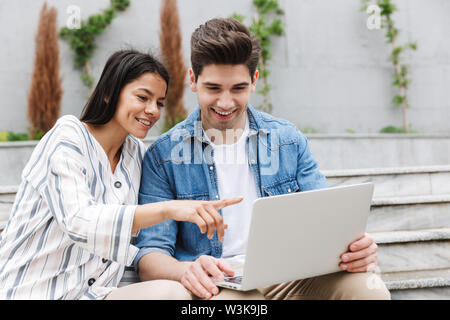 Immagine di carino coppia uomo e donna in abiti casual utilizzando e puntare il dito al computer portatile mentre è seduto sul banco vicino a scale all'aperto Foto Stock