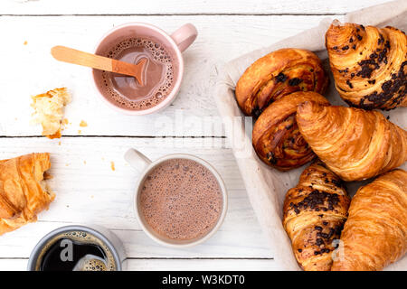 La prima colazione continentale con cioccolata calda, caffè nero e cesto di dolci. Mezza mangiata in legno bianco dal di sopra. Foto Stock