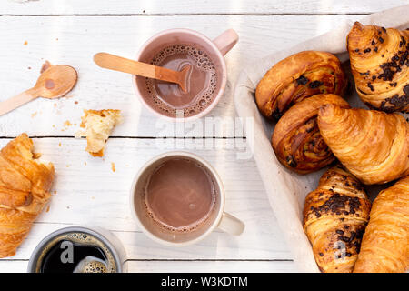 La prima colazione continentale con cioccolata calda, caffè nero e cesto di dolci. Mezza mangiata in legno bianco dal di sopra. Foto Stock