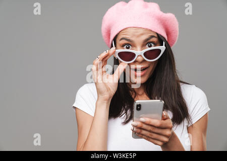 Photo closeup di stupito pretty woman in beret chiedendo e toccando i suoi occhiali da sole mentre si tiene lo smartphone isolate su uno sfondo grigio Foto Stock