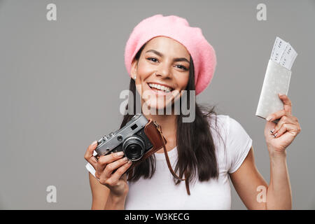 Foto Dettaglio di carino bella donna turistica in berretto di ridere mentre si tiene il retro della fotocamera e i biglietti di viaggio isolate su uno sfondo grigio Foto Stock