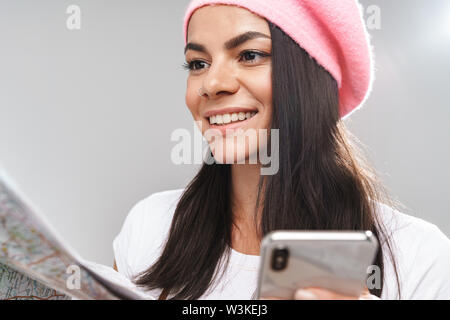 Primo piano immagine della cute giovane turista donna sorridente tenendo mappa su carta e cellulare isolate su uno sfondo grigio Foto Stock