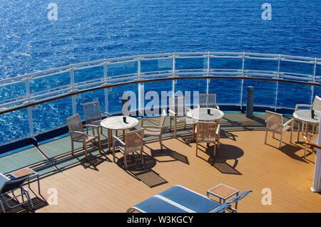 Bianco tavoli da pranzo con sedie e lettini per prendere il sole sul ponte della nave contro il blu oceano nel tempo soleggiato. Foto Stock