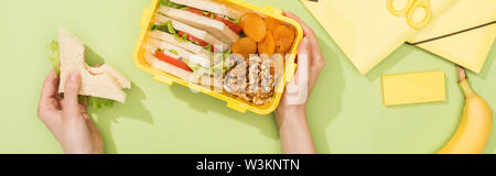 Vista ritagliata della donna azienda sandwich, scatola di pranzo con il cibo nei pressi di banana e articoli di cancelleria Foto Stock