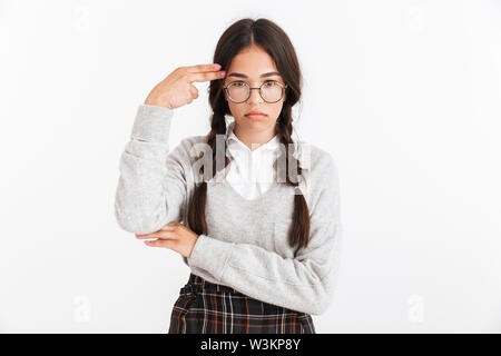 Foto Dettaglio di premuto ragazza adolescente indossando occhiali e uniformi scolastiche che mostra il dito il gesto della pistola al suo tempio isolate su sfondo bianco Foto Stock