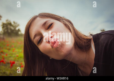 Giovane donna rendendo duckface kiss tenendo selfie foto con il suo smartphone o fotocamera nel campo di papaveri rossi. Ella divertirsi facendo sciocco e divertimento Foto Stock