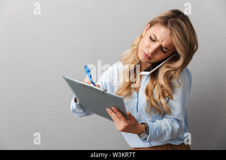 Foto di grave segretario bionda donna con lunghi capelli ricci a scrivere delle note in Appunti durante la chiamata sullo smartphone isolate su uno sfondo grigio Foto Stock