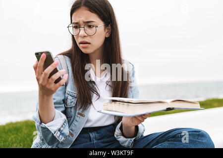 Immagine di un confuso orinato emotivo piuttosto giovane donna studente indossando occhiali passeggiate all'aperto libro lettura utilizzando il telefono cellulare. Foto Stock