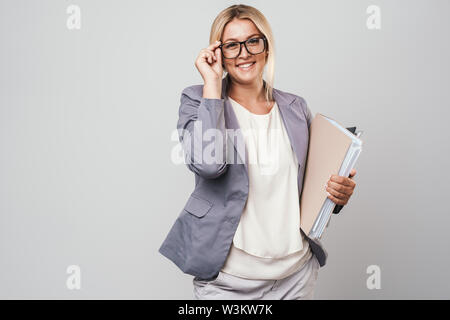 Attraente dai capelli biondi imprenditrice in giacca standing isolate su uno sfondo grigio, portando le cartelle Foto Stock