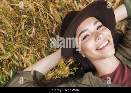 Foto dalla parte superiore della giovane donna con lunghi capelli scuri che indossa cappello elegante sorridente sdraiati sull'erba all'esterno Foto Stock