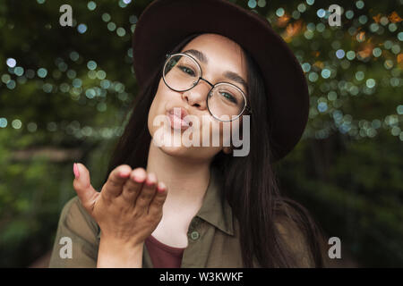 Foto Dettaglio di bella donna che indossa un cappello e occhiali di inviare aria kiss mentre passeggiate nel parco di sera Foto Stock